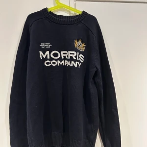 Svart stickad tröja från Morris - Säljer en stilren svart stickad tröja från Morris med vit text och ett broderat emblem på bröstet. 