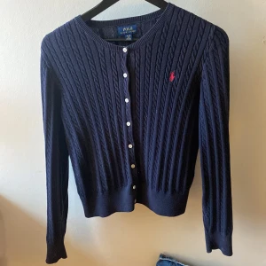 Mörkblå kofta från Ralph Lauren - Säljer en stilren mörkblå kofta från Ralph Lauren med kabelstickat mönster och knappar framtill. Den har en klassisk design med långärmade ärmar och en liten röd logotyp på bröstet. Perfekt för en elegant och avslappnad look.