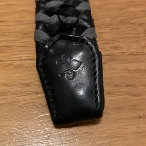 Morris flättat bälte  - Snyggt flätat bälte i svart och grått med en stilren silverfärgad spänne. Bältet har en unik design med ett flätat mönster och en elegant detalj i skinn vid änden. Perfekt för att ge en extra touch till din outfit.