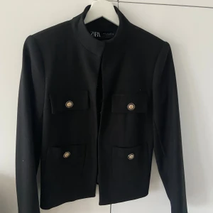 Svart kavaj från Zara - Elegant svart kavaj från Zara med långa ärmar och stilrena knappar framtill. Kavajen har en klassisk design med två fickor på bröstet och en rak passform. Perfekt för en stilren look.