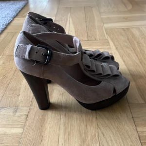 Beige klackskor från Topshop - Snygga beige klackskor från Topshop i mocka med volangdetaljer och öppen tå. Skorna har en rejäl klack och justerbart spänne vid ankeln för perfekt passform.