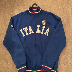 Italia Crewneck - Sitter som L/XL. Dvs är taggen missledande.Säljer en blå jacka med texten 'ITALIA' och Italiens flagga på bröstet och ärmen. Jackan har dragkedja och ribbade muddar i grönt, vitt och rött. Perfekt för att visa ditt stöd för Italien!