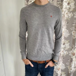 Grå tröja i merinoull från Morris - Säljer en stilren grå tröja från Morris i 100% merinoull. Tröjan har en liten broderad logotyp på bröstet och ribbade muddar vid ärmslut och nederkant. Perfekt för en elegant och bekväm look.