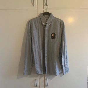 Grå skjorta från A Bathing Ape - Snygg grå skjorta från A Bathing Ape med ikonisk logga på bröstet. Skjortan har långa ärmar och knappar framtill. Perfekt för dig som gillar streetwear med en touch av stil. Tröjan är i Strl M men passar M/S