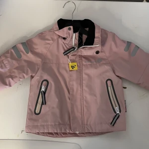 Rosa jacka från On The Peak - Säljer en söt rosa jacka storlek 80 från On The Peak med reflexdetaljer på ärmarna och dragkedjor på fickorna. Jackan har en hög krage och knappar vid halsen för extra skydd mot väder. Perfekt för små äventyrare!