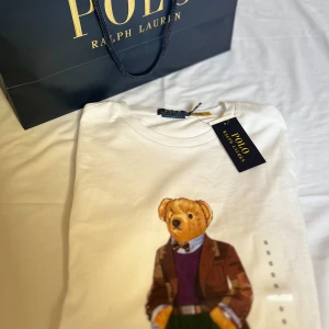 Polo Bear T-shirt från Ralph Lauren - Säljer en vit t-shirt från Ralph Lauren med den ikoniska Polo Bear tryckt på framsidan. Björnen är klädd i en stilfull outfit med kavaj och tröja. T-shirten har en rund hals och är kortärmad, perfekt för en avslappnad stil. Kvitto ingår. Nypris 1199. Bara att höra av sig vid frågor//Carl