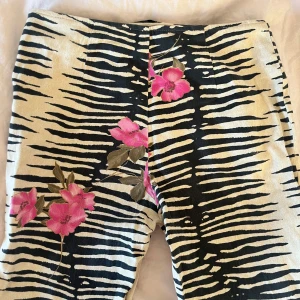 Mönstrade byxor med blommor - Snygga byxor med ett djärvt zebramönster i svart och beige, dekorerade med rosa blommor. Byxorna har en dragkedja för enkel på- och avtagning. Passar M-L