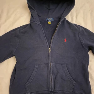 Mörkblå hoodie från Ralph Lauren - Säljer en snygg mörkblå hoodie från Ralph Lauren med dragkedja och huva. Den har en röd broderad logga på bröstet och praktiska fickor framtill. Perfekt för en avslappnad stil. Mycket bra skick 9/10