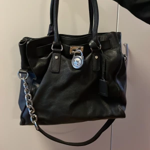 Svart handväska med kedjedetalj från Michael Kors - Snygg svart handväska i skinn med silverfärgad kedjedetalj och lås framtill. Väskan har två handtag och en rymlig insida med flera fack. Perfekt för att bära med sig alla nödvändigheter med stil.