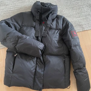 Canada Snow Monroe Puffer jacket - Säljer en svart dunjacka från Canada Snow med hög krage och dragkedja. Jackan har långa ärmar och en logotyp på ärmen. Perfekt för kalla dagar.