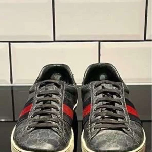 Äkta Gucci Ace Skor - Snygga svarta sneakers med ett diskret mönster och ikoniska ränder i rött och blått på sidan. Skorna har snörning och en vit sula som ger en klassisk look. Perfekta för en stilren vardagsstil.