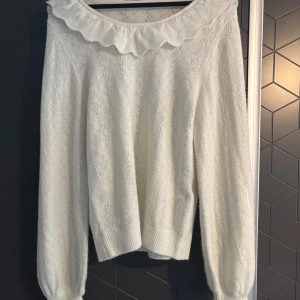 Vit stickad tröja med volangdetalj - Säljer en söt vit stickad tröja med volangdetalj runt halsen. Tröjan har långa ärmar och en mjuk, bekväm passform. Perfekt för kyligare dagar.