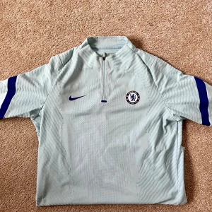 Ljusblå träningströja från Nike - Säljer en ljusblå träningströja från Nike med Chelsea FC-logga på bröstet. Tröjan har en halv dragkedja och mörkblå detaljer på ärmarna. Perfekt för fotbollsträning eller som en sportig vardagströja.