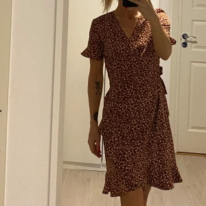 Blommig omlottklänning från Only - Säljer en söt blommig omlottklänning i rödbrun nyans med korta ärmar och volangdetaljer. Perfekt för en avslappnad stil med sneakers eller klä upp med klackar. Klänningen har en v-ringad hals och knytning i midjan för en smickrande passform.
