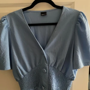 Blå blus från Gina Tricot - Säljer en söt blå blus från Gina Tricot med korta ärmar och knappar framtill. Blusen har en smockad midja som ger en fin passform. Perfekt för vår och sommar! Stlk L men liten i storlek så passar både S och M