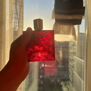 Röd parfymflaska från Versace - Snygg röd parfymflaska från Versace med ett elegant mönster och en stilren svart kork. Flaskan har en fyrkantig form och ger en lyxig känsla. Perfekt för den som vill ha en doft med attityd och stil.