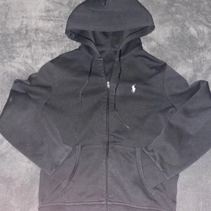 Svart hoodie från Polo - Säljer en stilren svart hoodie från Polo med dragkedja och klassisk logga på bröstet. Tröjan har en bekväm passform och praktiska fickor framtill. Perfekt för en avslappnad look.