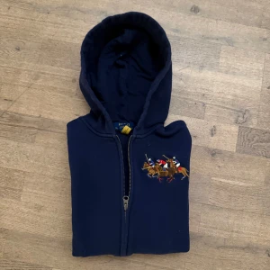  Polo Ralph Lauren tröja 164 - Snygg mörkblå hoodie från Polo Ralph Lauren med dragkedja och broderad polospelare på bröstet. Tröjan har långa ärmar och en praktisk huva. Perfekt för en avslappnad stil.