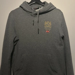 Grå hoodie från Morris - Säljer en stilren grå hoodie från Morris med ett broderat emblem och brittisk flagga på bröstet. Tröjan har en klassisk känguruficka och justerbar huva med snören. Perfekt för en avslappnad stil.