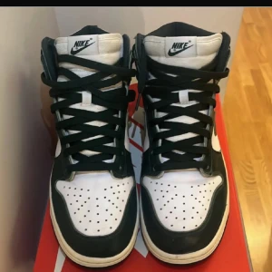 Nike Dunk High Spartan Green - Snygga Nike Dunk High i färgen Spartan Green. Skorna har en klassisk design med vit och grön färgkombination. De har snörning och en hög siluett som ger ett sportigt utseende. Perfekta för sneakerälskare som vill ha en stilren look.