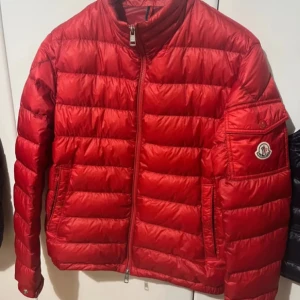 Röd pufferjacka från Moncler - Säljer en snygg röd pufferjacka från Moncler. Jackan har en dragkedja framtill och ett broderat Moncler-märke på ärmen. Perfekt för kyliga dagar med sin quiltade design.