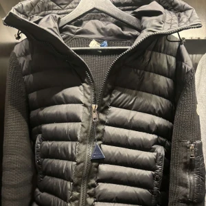 Svart jacka från Moncler - Säljer en snygg svart jacka från Moncler med quiltad design och dragkedja. Jackan har en huva och ärmar i stickat material, vilket ger en cool kontrast. Perfekt för kyligare dagar! Liten hål som syns på bilden utöver det inga defekter.
