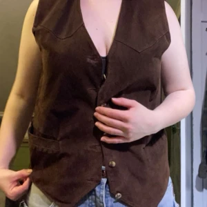Brun mockaväst med knappar - Säljer en brun väst med knappar framtill. Västen har en v-ringad design och två fickor. Knapparna är troligtvis påsydda för hand och sitter lite lösa men lätt att fixa.
