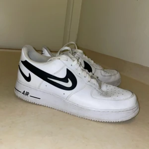 Nike Air Force 1 i vitt och svart - Säljer ett par klassiska Nike Air Force 1 sneakers i vitt med svarta detaljer. Skorna har snörning och den ikoniska Nike-loggan på sidan. Perfekta för en stilren look. Skorna är hela men behöver sig en noggrannare rengöring 