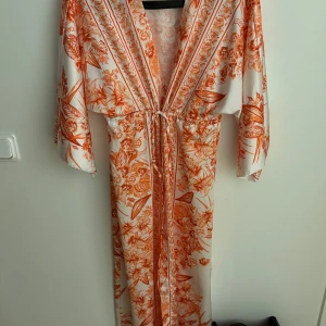 Kimono - Säljer en orange Kimono med vita blommönster kimonon har trekvartsärmar och ett knytband i midjan för en justerbar passform. Perfekt för somriga dagar eller speciella tillfällen. Inte använt då jag upplever färgen inte passade mig. Kimonon är beställd från Frankrike så jag orkade inte returnera tillbaka den. 