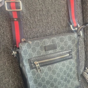 Grå axelväska från Gucci - Snygg grå axelväska från Gucci med klassiskt mönster och justerbar axelrem i rött och blått. Väskan har en praktisk dragkedja framtill och är perfekt för att bära med sig det viktigaste. En stilren accessoar för alla tillfällen.Den är använd därför priset är lågt.