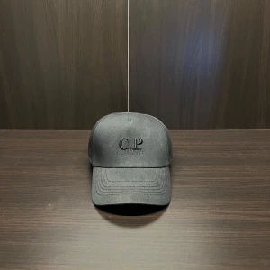 Svart keps från C.P. Company - Snygg svart keps från C.P. Company med broderad logga framtill. Kepsen har en unik design med goggle baktill, vilket ger den en cool och annorlunda look. Perfekt för dig som vill sticka ut!