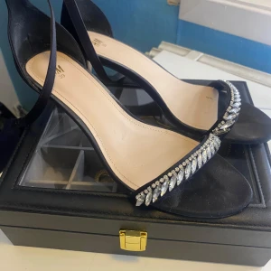 Svarta klackskor med strassdetaljer från H&M - Eleganta svarta klackskor från H&M med glittrande strassdetaljer över tårna. Skorna har en smal rem runt ankeln och en stilren design som passar perfekt för festliga tillfällen. Enbart används under balen🤍🤍