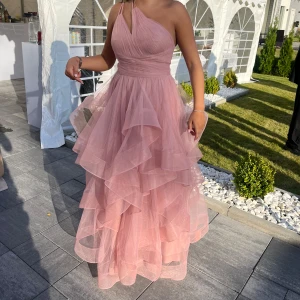 Rosa långklänning med volanger - Elegant rosa långklänning med volanger och en axelbandslös design. Klänningen har en vacker omlottstil och en subtil slits framtill. Perfekt för speciella tillfällen som bal eller bröllop. Originalpris 3000kr