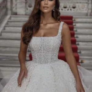 Vit brudklänning med pärlor - Elegant vit brudklänning med pärlor och breda axelband. Klänningen har en klassisk lång design med en fyllig kjol och ett vackert mönster av pärlor över hela tyget. Perfekt för en sagolik bröllopsdag.