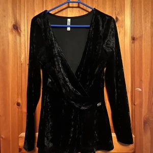 Svart sammetstopp från Chiara Forati Milano - Elegant svart sammetstopp från Chiara Forati Milano med omlott-design och långa ärmar. Perfekt för en stilfull look med en mjuk och lyxig känsla. Toppen har en v-ringad hals och en dekorativ detalj vid midjan.