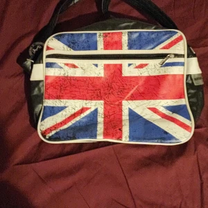 Axelväska med Union Jack från Robin Ruth - Snygg axelväska från Robin Ruth med Union Jack-mönster på framsidan. Väskan har en dragkedja och är tillverkad i syntetmaterial. Perfekt för dig som vill ha en unik och stilren väska med brittisk touch.