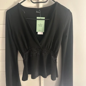 Svart omlottopp med spetsdetaljer från Gina Tricot - Snygg svart omlottopp från Gina Tricot med spetsdetaljer runt midjan. Toppen har långa ärmar och en elegant v-ringning. Perfekt för att ge en stilren look. Strl XXS. Lapparna sitter kvar🩷🩷