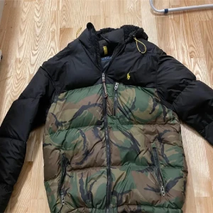 Camouflage pufferjacka från Ralph Lauren - Säljer en snygg pufferjacka från Ralph Lauren i camouflage och svart. Jackan har en dragkedja framtill och två praktiska fickor med dragkedjor. Perfekt för höst och vinter, håller dig varm och stilren. Den har långa ärmar och en liten broderad logga på bröstet. Passar perfekt för både vardag och äventyret !