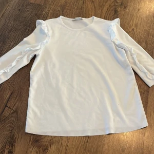 Vit långärmad topp med volangdetaljer - Säljer en superfin vit topp med långa ärmar och volangdetaljer längs axlarna. Perfekt för både vardag och fest! Materialet känns mjukt och bekvämt, och den har en normal passform. Passar perfekt till jeans eller kjol för en stilren look.