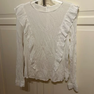 Vit långärmad blus med volangdetaljer - Säljer en söt vit blus från Kappahl i storlek S. Den är gjord av 100% bomull och har långa ärmar med fina volangdetaljer längs framsidan och ärmarna. Perfekt för både vardag och festliga tillfällen. Blusen har en dragkedja i ryggen för enkel påklädning.