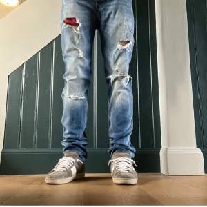 Säljer ett par blå jeans med slitningar och hål för en cool och avslappnad look. De har en slim passform och är perfekta för vardagsbruk. Jeansen har en klassisk femficksdesign och är tillverkade i denim. Perfekta för både höst och vår!