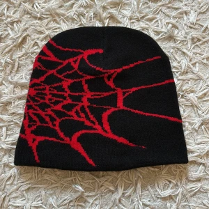 Svart mössa med rött spindelnätsmönster - Cool svart mössa med ett rött spindelnätsmönster. Perfekt för att ge din outfit en edgy touch. Den är mjuk och bekväm, idealisk för kyliga dagar. Passar bra till både vardag och speciella tillfällen. Helt ny, oanvänd mjuk bomullsmössa, passar perfekt för streetwear och y2k. Hör av dig vid minsta intresse!