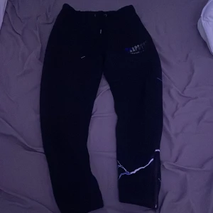 Svarta Trapstar mjukisbyxor - Säljer ett par svarta mjukisbyxor från Trapstar med ett coolt blixtmönster i vitt och blått längs ena benet. Byxorna har en bekväm passform och är perfekta för chill dagar. De har även en elastisk midja med snörning för bästa komfort. Perfekta för både vardag och avslappnade tillfällen!