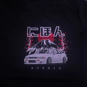 Svart t-shirt med japanskt motiv - Snygg svart t-shirt med ett coolt japanskt motiv. Trycket visar en bil framför ett berg med en röd sol i bakgrunden och japanska tecken. Perfekt för bilentusiaster och streetwear-stil. Passar bra till både vardag och fest!