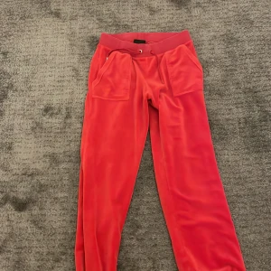 Röda mjukisbyxor från Juicy Couture - Säljer ett par snygga röda mjukisbyxor från Juicy Couture i storlek S. Byxorna är i mjukt material med elastisk midja och snörning. De har praktiska fickor med knappar bak och en broderad logga framtill. Perfekta för en avslappnad stil!