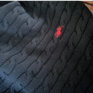 Mörkblå stickad tröja från Ralph Lauren - Säljer en snygg mörkblå stickad tröja från Ralph Lauren med det klassiska röda logotypbroderiet på bröstet. Tröjan har en kabelstickad design och långa ärmar med ribbade muddar. Perfekt för både höst och vinter!