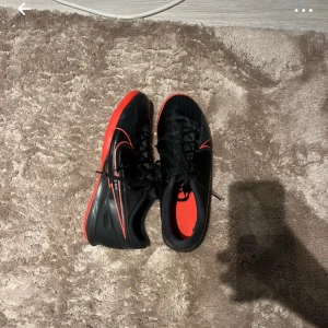 Svarta och röda fotbollsskor - Säljer ett par snygga svarta fotbollsskor med röda detaljer. Skorna har snörning och en cool design med en röd sula som ger extra grepp på planen. Perfekta för fotbollsträning eller match!