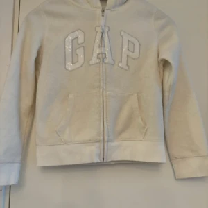 Vit hoodie från GapKids - Säljer en supermjuk och skön vit hoodie från GapKids. Den har en dragkedja framtill och stora fickor. På bröstet står det 'GAP' med glittriga bokstäver. Perfekt för kyliga dagar eller mysiga stunder hemma. Passar barn i åldern 10-11 år. Har en lite blå fläck längst ner på vänster arm (de sista bilden), inget man tänker på, men annars bra skick. 