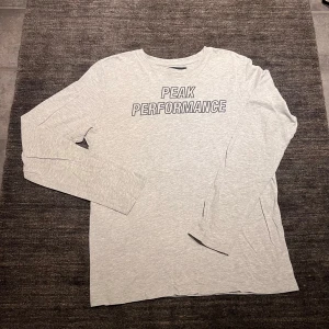 Grå långärmad tröja från Peak Performance - Säljer en grå långärmad tröja från Peak Performance. Den har en stilren design med text på framsidan. Tyvärr finns det ett litet hål på ärmen, men annars i bra skick. Perfekt för vardagsbruk eller träning!