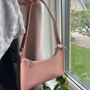 Rosa axelväska i syntet - Snygg rosa axelväska i syntetmaterial med krokodilpräglat mönster. Väskan har en enkel design med en dragkedja och en liten dekorativ knappdetalj. Perfekt för både vardag och fest!